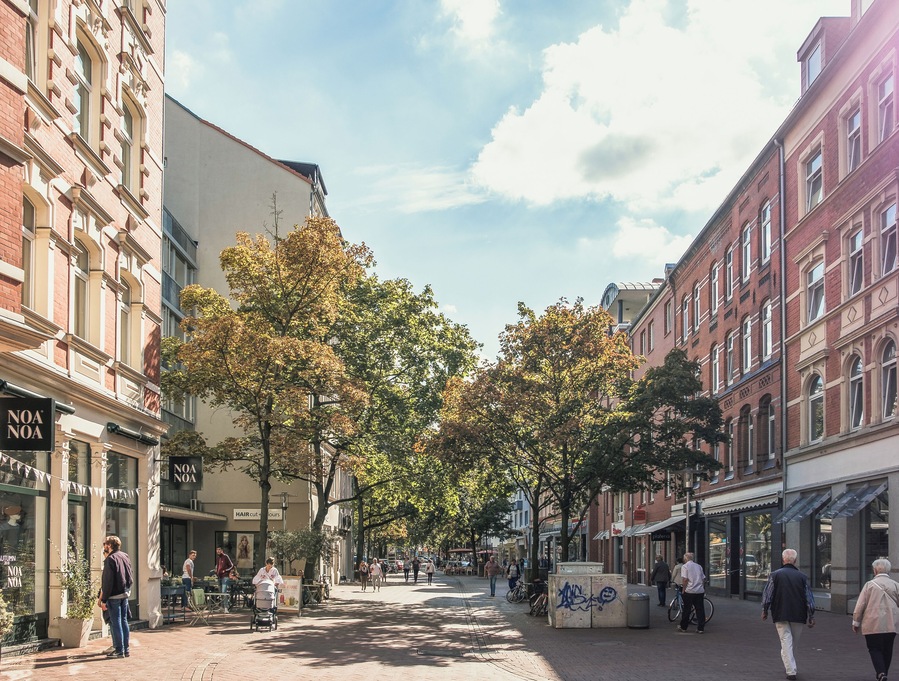 A tree-lined cobblestone street with historic buildings, a café, and pedestrians.