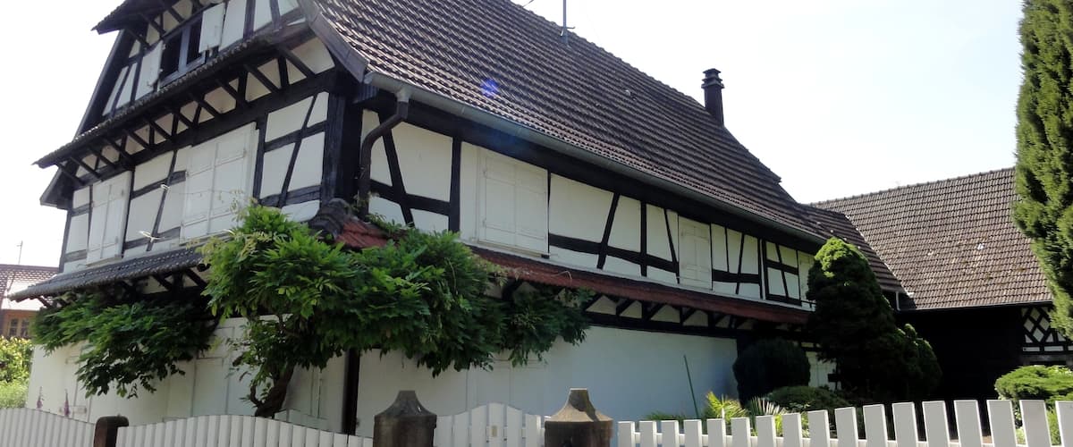 A traditional half-timbered house with a steeply pitched roof, surrounded by a white picket fence and lush greenery.