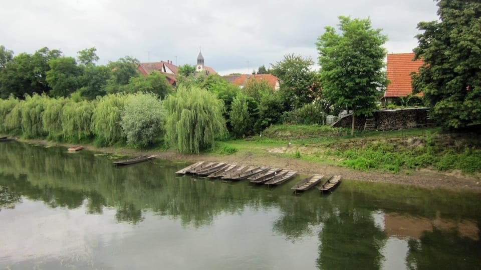 A calm river with wooden boats docked on the bank, surrounded by lush greenery and traditional buildings with red roofs.
