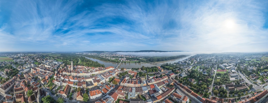 A panoramic view of a town with a river, surrounded by greenery and a clear sky.