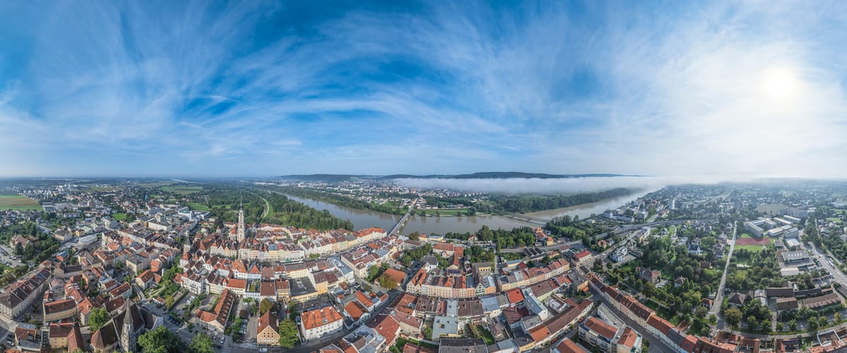 A panoramic view of a town with a river, surrounded by greenery and a clear sky.