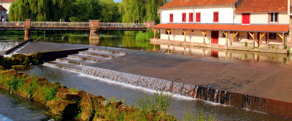A river with a small waterfall, a bridge, and buildings with red shutters.