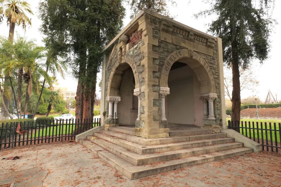 A stone structure with arches and columns, labeled "Public Library," surrounded by trees and a fenced area.