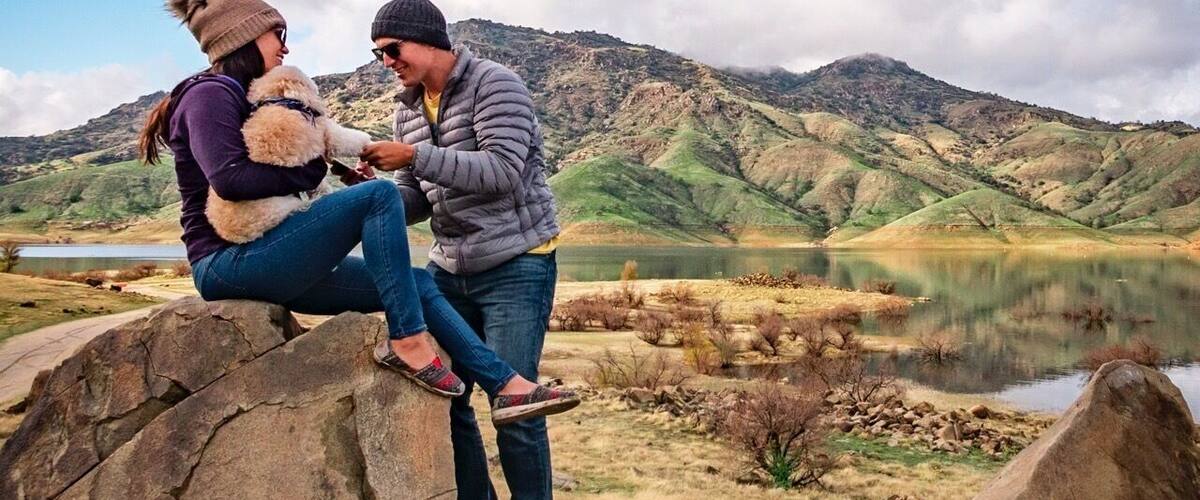 Two people sitting on rocks by a lake with mountains in the background.