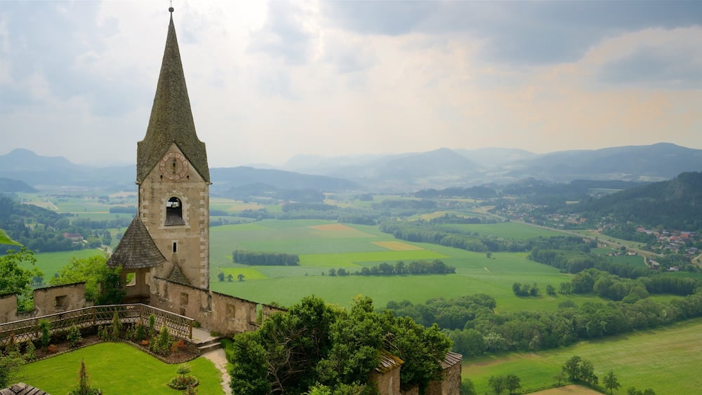 A church with a tall spire and a clock tower, surrounded by lush green fields and rolling hills in the background.