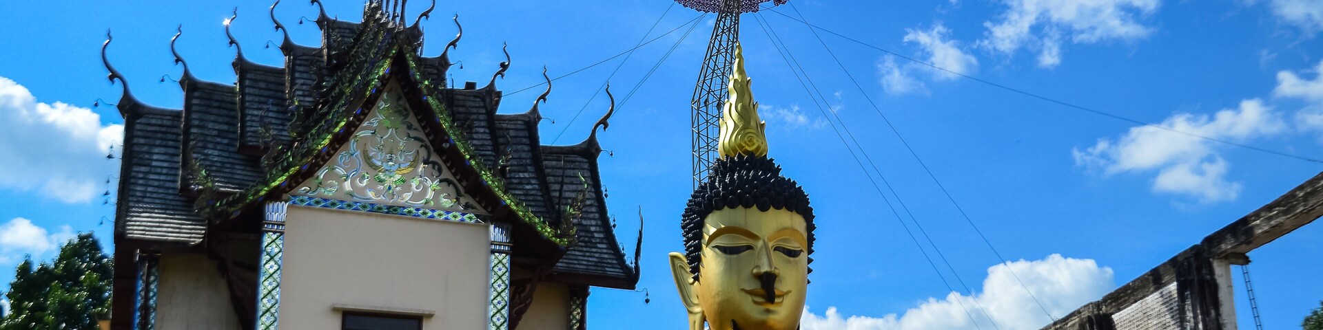 Una estatua dorada y grande de Buda con una parte superior puntiaguda, un edificio tradicional con tejado de tejas y un cielo azul claro con nubes.