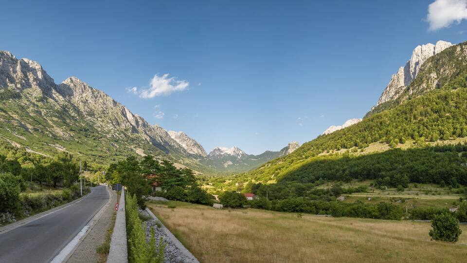 A mountain road flanked by lush green hills and rocky peaks under a clear blue sky.