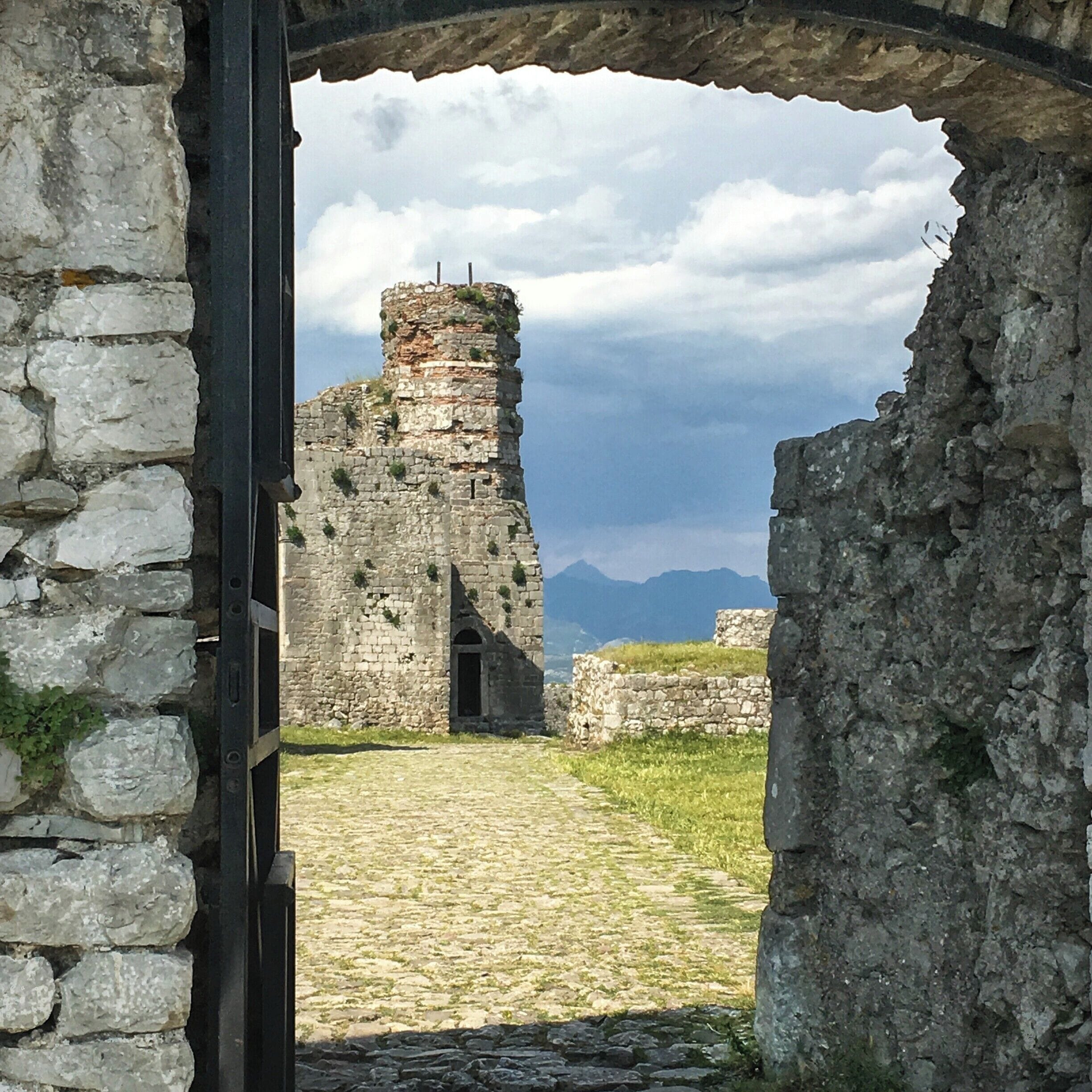 An ancient stone structure with a tower, surrounded by grassy terrain and mountains in the background.