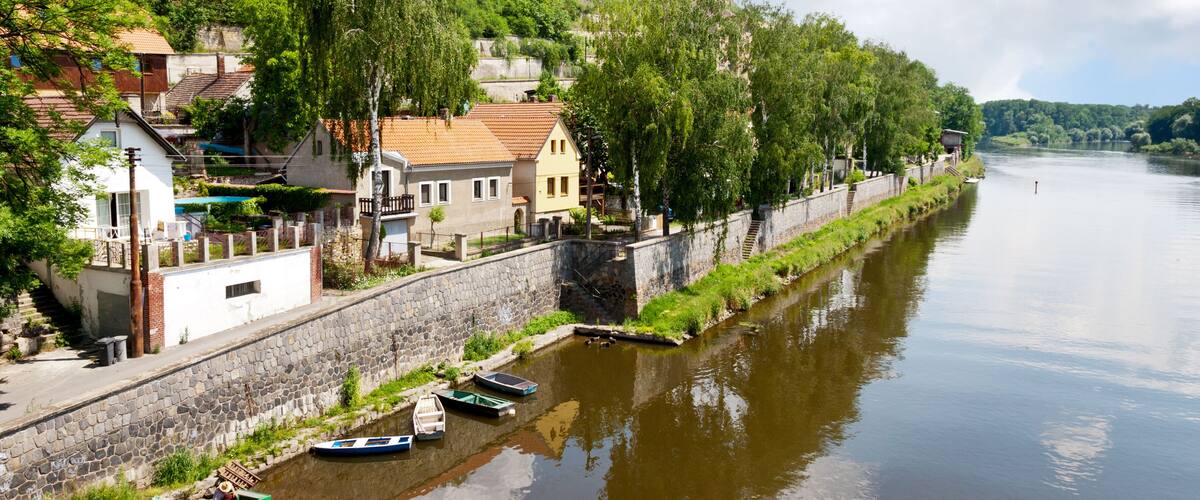 A river with boats, a stone wall, and buildings on a hillside.