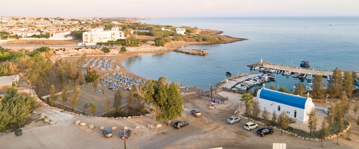 Aerial bird's eye view of coastline sunset, landmark white washed chapel Agia Triada beach, Protaras, Famagusta, Cyprus from above. Tourist attraction Ayia Trias bay church, sand, port at sunrise.