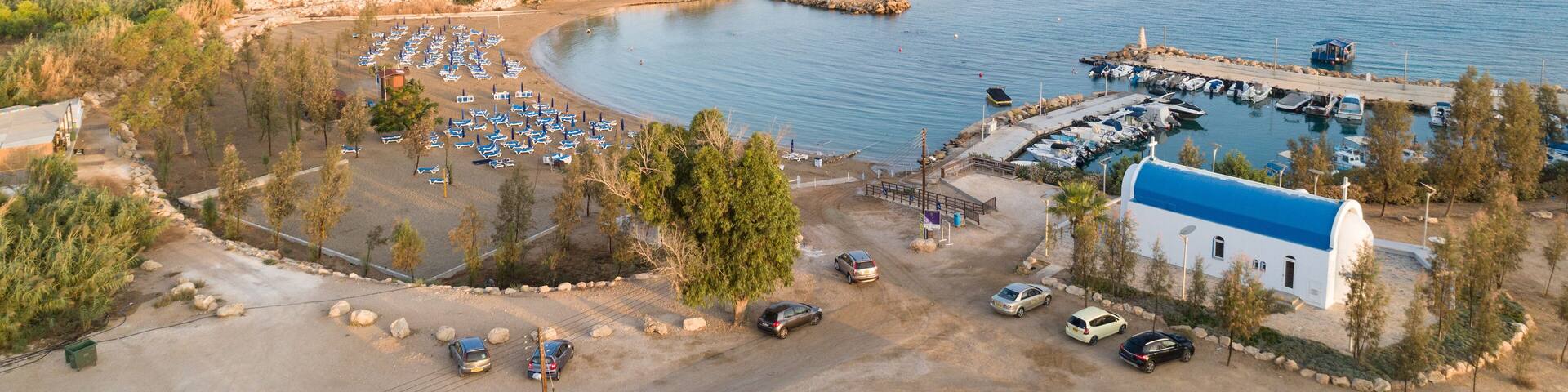 Aerial bird's eye view of coastline sunset, landmark white washed chapel Agia Triada beach, Protaras, Famagusta, Cyprus from above. Tourist attraction Ayia Trias bay church, sand, port at sunrise.