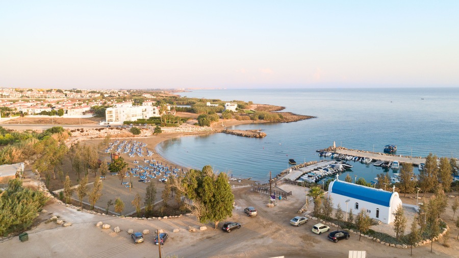 Aerial bird's eye view of coastline sunset, landmark white washed chapel Agia Triada beach, Protaras, Famagusta, Cyprus from above. Tourist attraction Ayia Trias bay church, sand, port at sunrise.