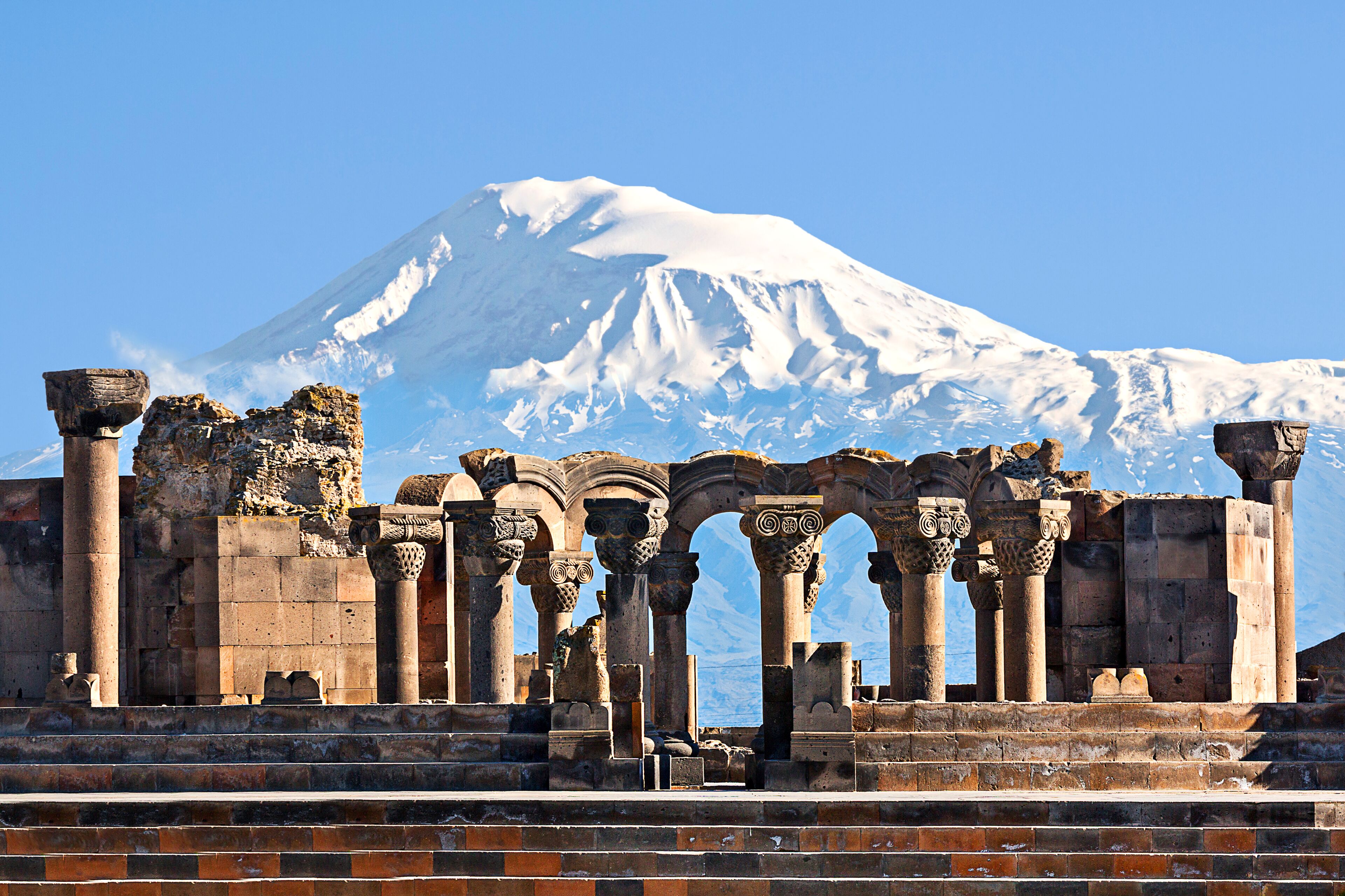 Ancient ruins with columns and arches in front of a snow-capped mountain.