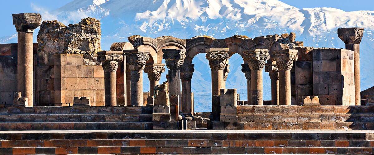 Ancient ruins with columns and arches in front of a snow-capped mountain.