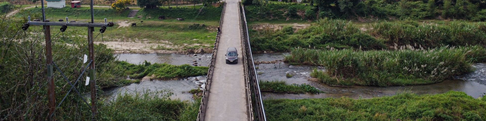 A car crossing a rural bridge over a canal surrounded by lush greenery and mountains in Ban Na San District, Surat Thani Province, Thailand.