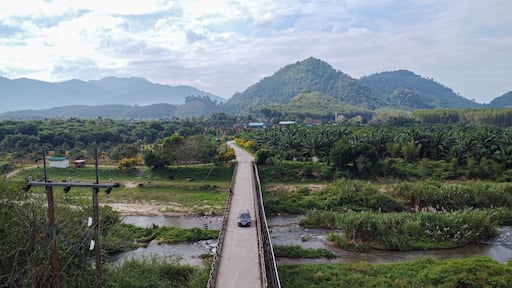 A car crossing a rural bridge over a canal surrounded by lush greenery and mountains in Ban Na San District, Surat Thani Province, Thailand.