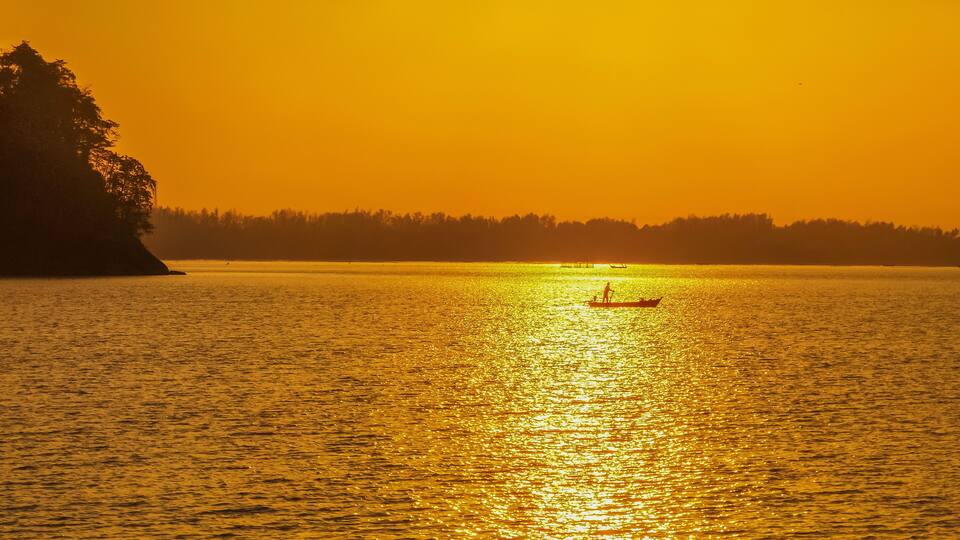 A sunset over a calm body of water with a silhouette of a large rock on the left and a small boat with a person in the distance.