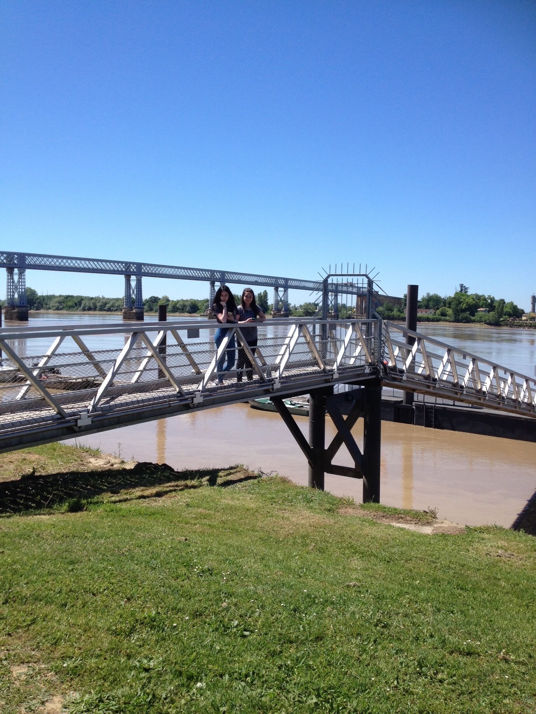 A pedestrian bridge over a river with a metal railing and a grassy bank in the foreground.