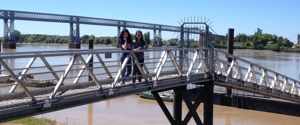 A pedestrian bridge over a river with a metal railing and a grassy bank in the foreground.