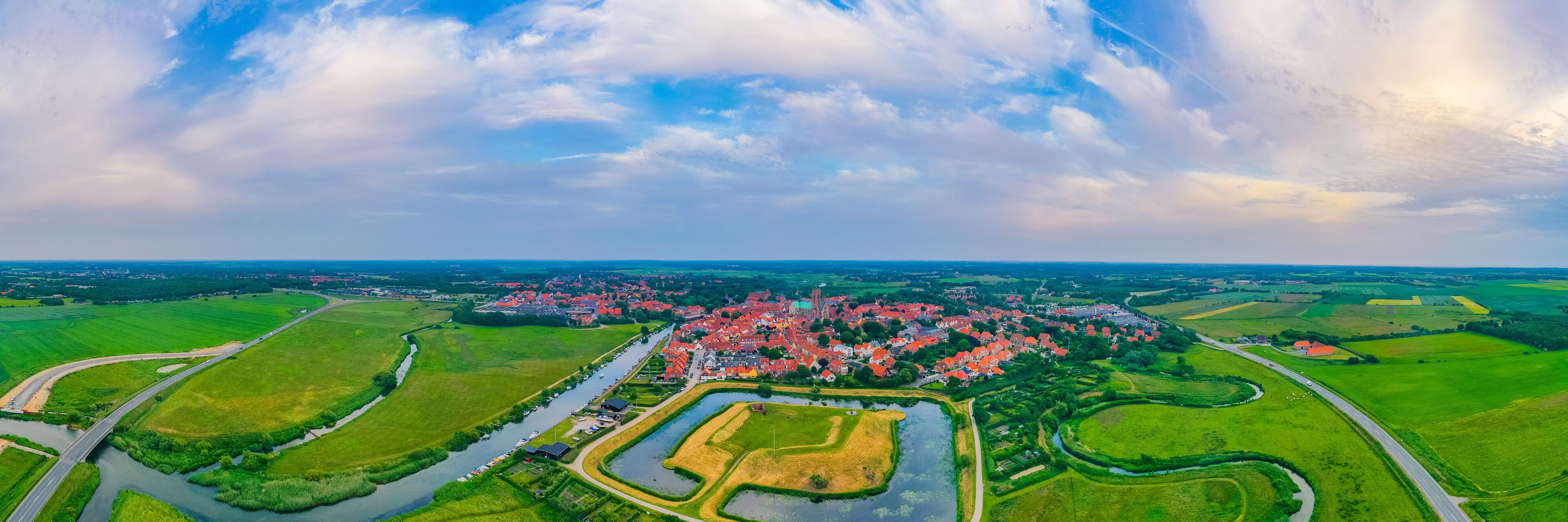 A town with red-roofed houses, surrounded by green fields and a river.