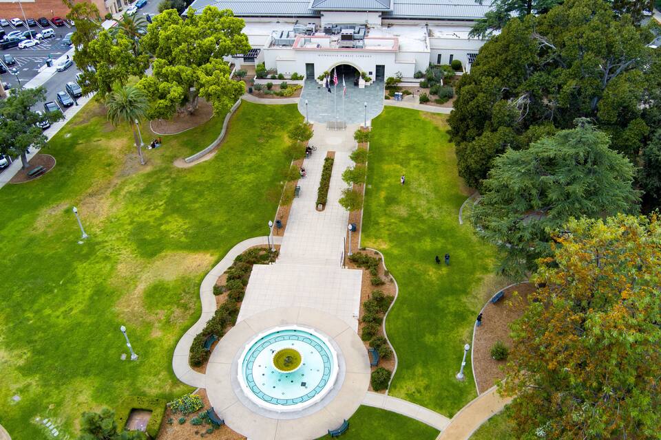 An aerial view of a landscaped area with a central fountain, surrounded by well-maintained lawns and trees.