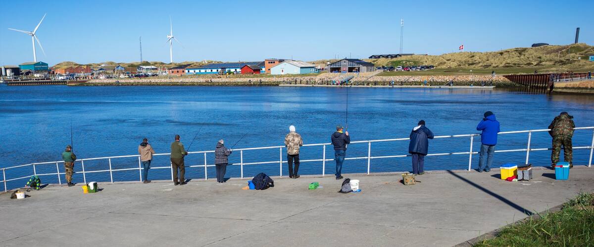 A waterfront with people fishing, wind turbines, and industrial buildings.