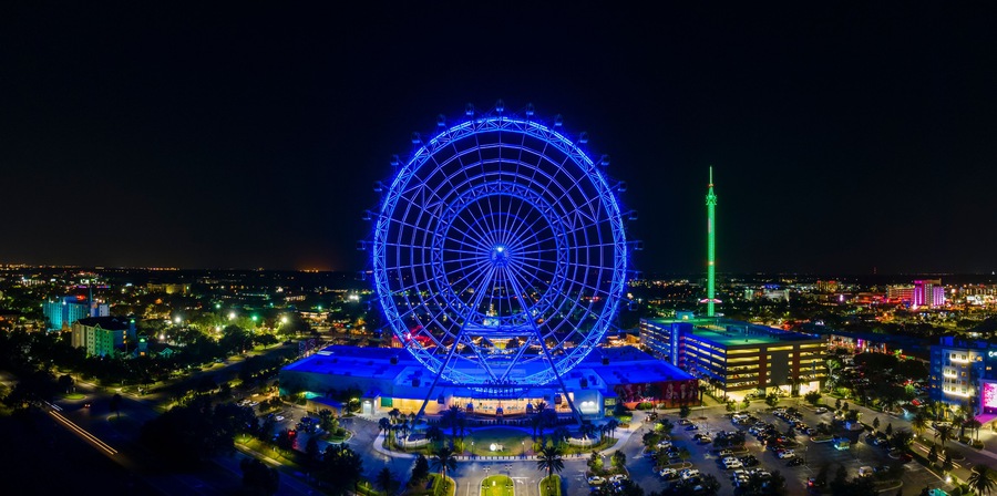 A large Ferris wheel with blue lights, surrounded by a cityscape with illuminated buildings and a tall green tower.