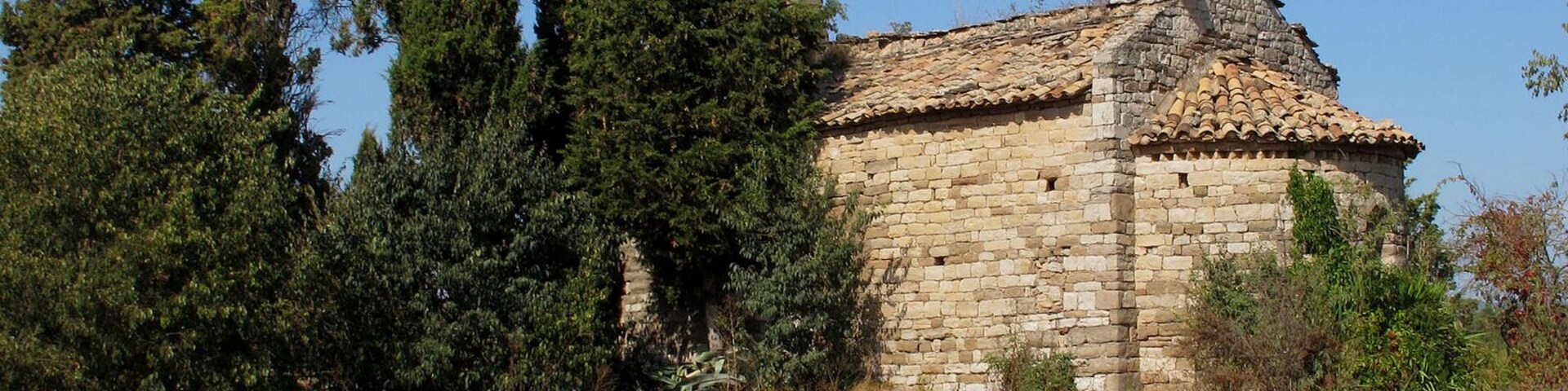 Um edifício de pedra com telhado de telhas, rodeado por árvores altas e um campo seco.