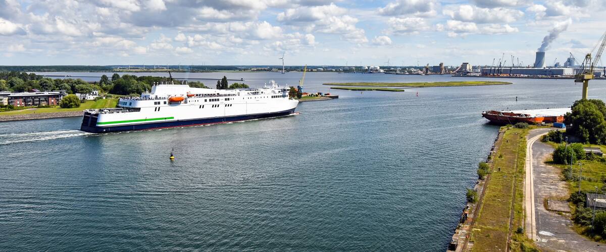 A large white ferry with green stripes on a river, surrounded by industrial buildings and a bridge in the background.