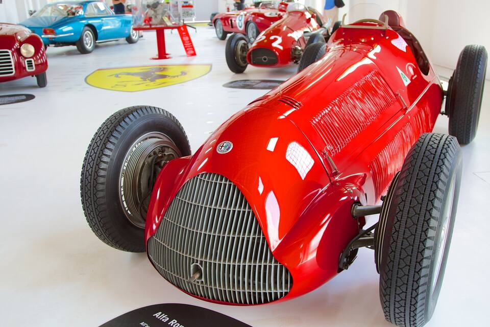 A red vintage race car with a sidecar, displayed in a museum with other classic cars.