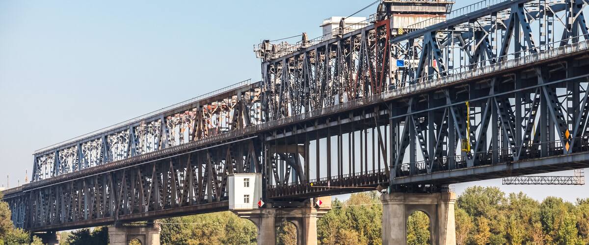 A large metal bridge spanning a river with a forested area in the background.