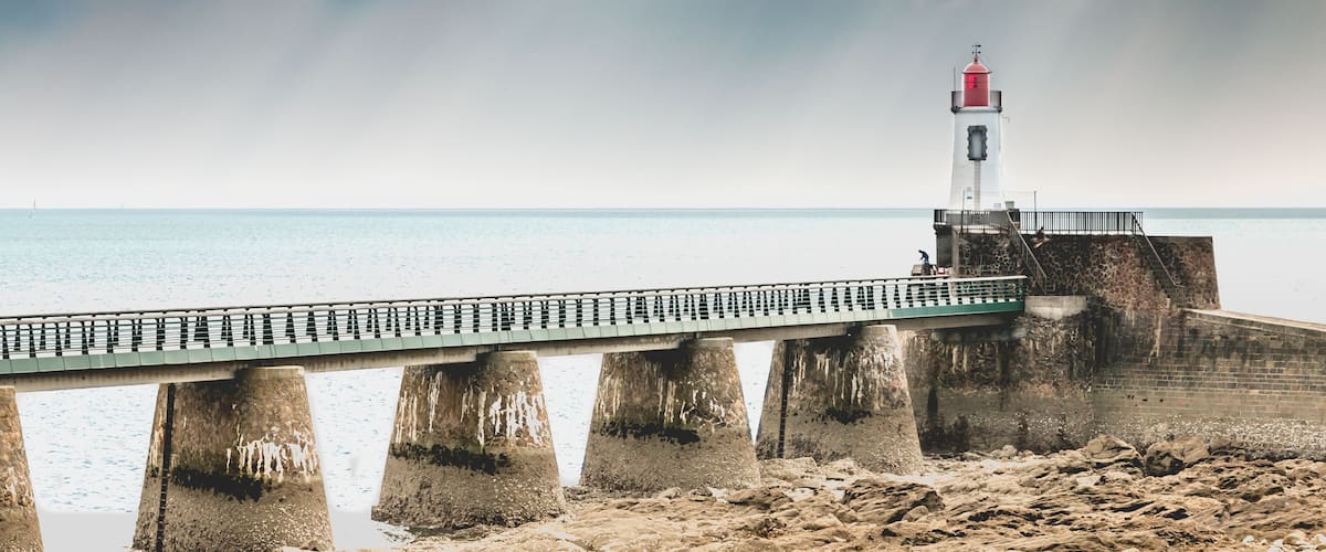 A lighthouse on a pier extending into the sea, with a rocky shoreline in the foreground.