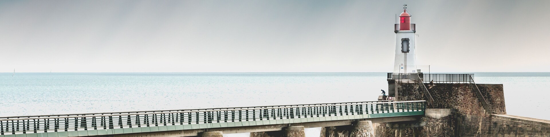 A lighthouse on a pier extending into the sea, with a rocky shoreline in the foreground.