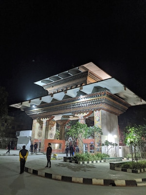 A brightly lit, intricately designed pavilion with ornate patterns and columns, surrounded by a paved area with plants and a few people.