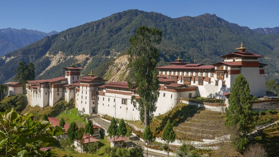 A traditional monastery complex with multiple buildings, surrounded by lush greenery and mountains.
