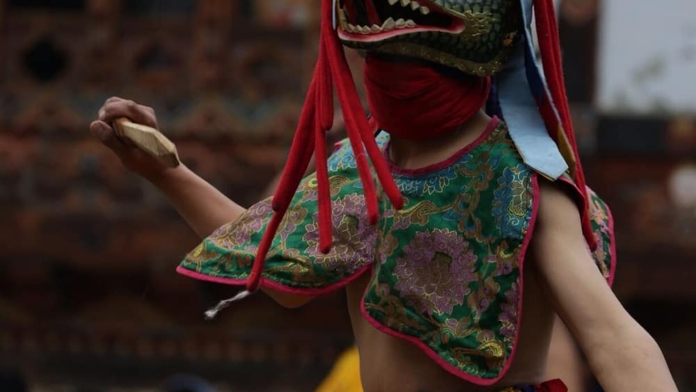 A person in traditional attire with a mask, colorful clothing, and a yellow skirt.