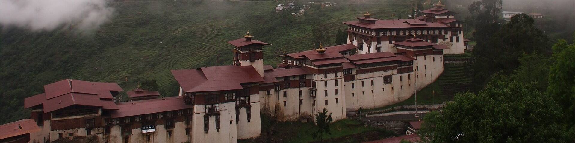 A monastery complex nestled in a valley with terraced fields and mist-covered mountains in the background.