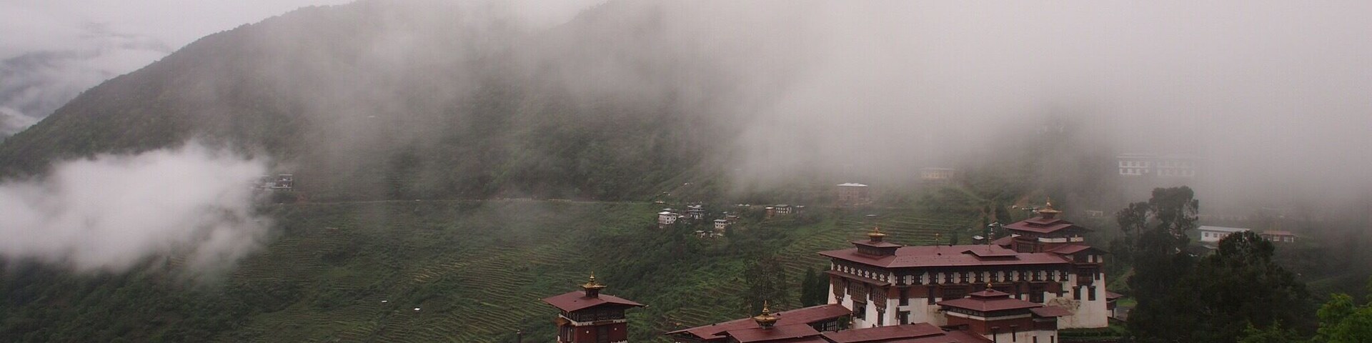 A monastery complex nestled in a valley with terraced fields and mist-covered mountains in the background.