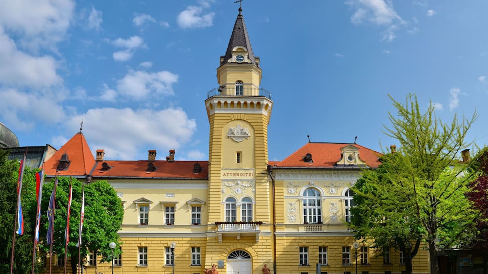 A historic yellow building with a clock tower, surrounded by flags and trees.
