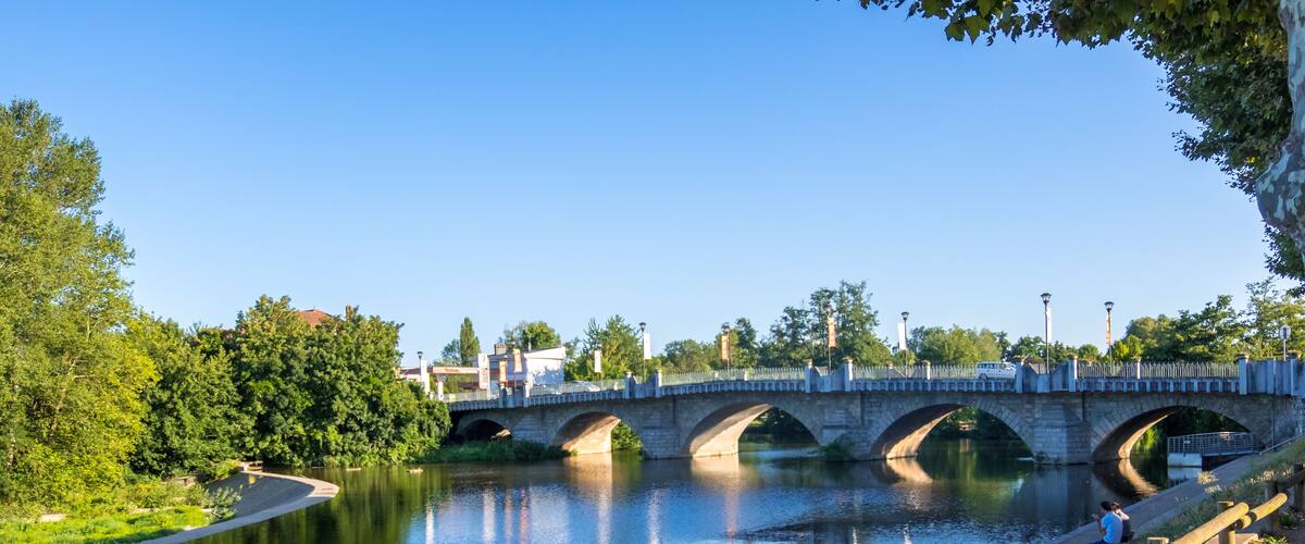 A stone bridge over a calm river, surrounded by greenery and trees.