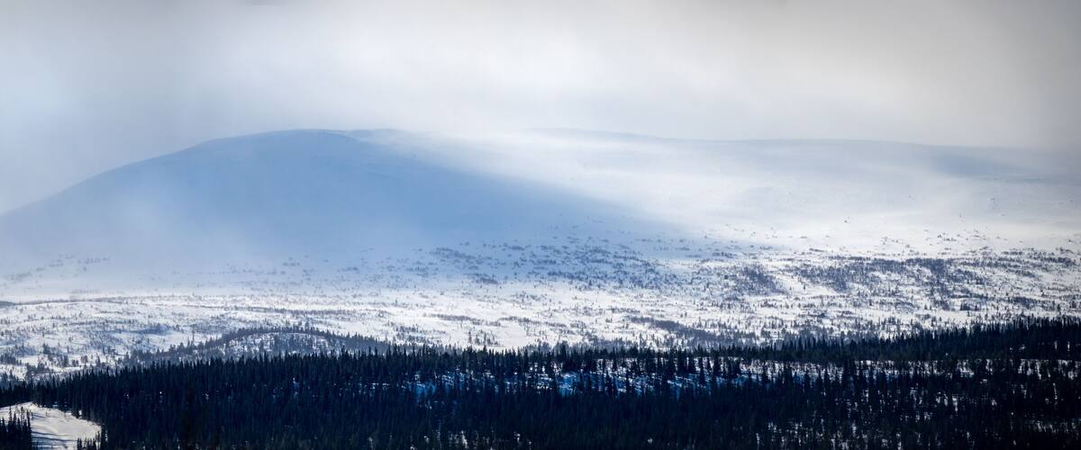 A snow-covered mountain with a forested foreground and a small building at the base.