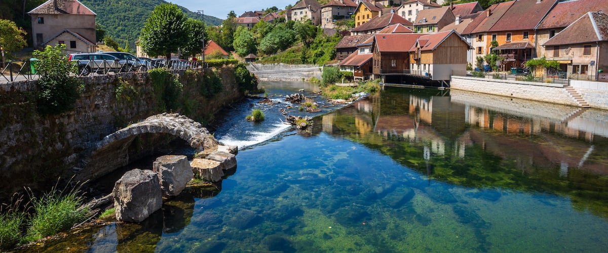 A clear river with a stone bridge, surrounded by houses and a church on a hillside.