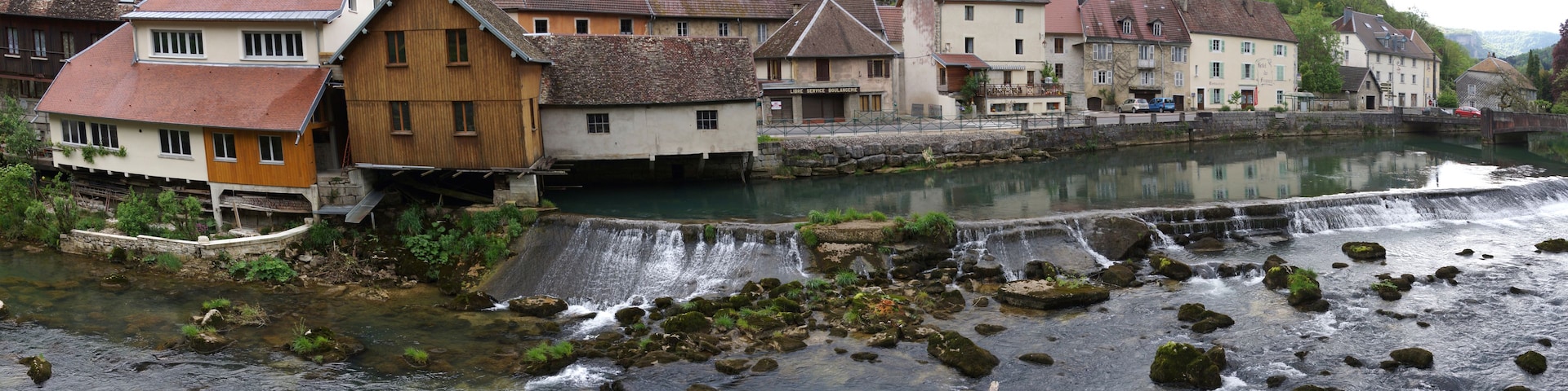 Une rivière avec une cascade, entourée de bâtiments traditionnels et d’une végétation luxuriante.