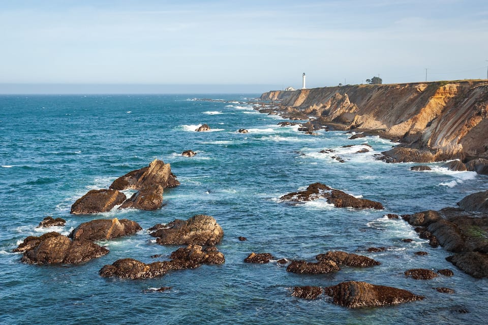 A rocky coastline with waves crashing against the rocks and a lighthouse in the distance.