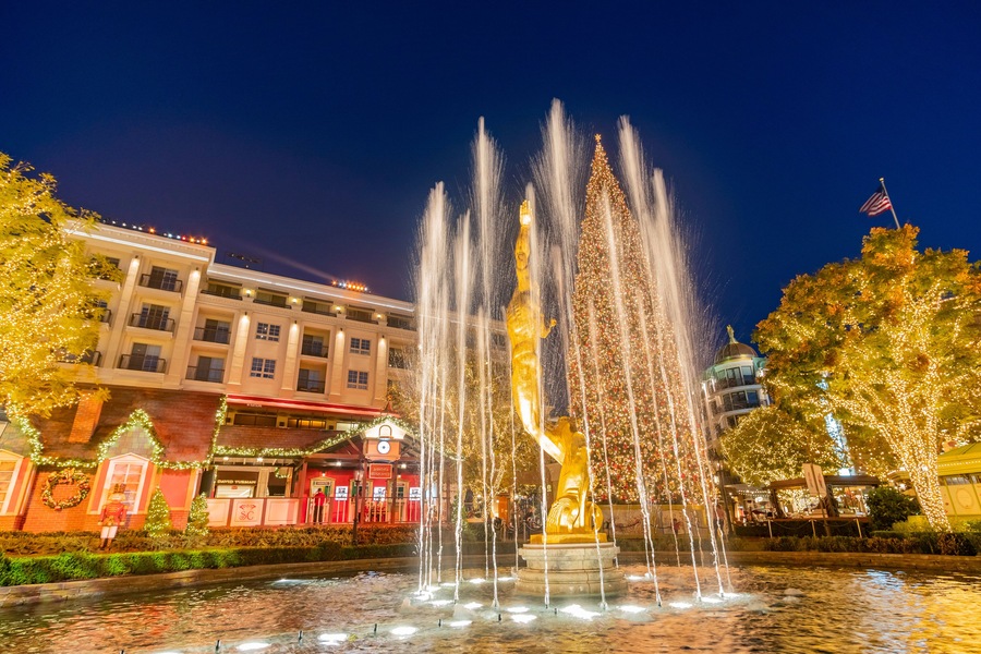 Una fuente iluminada con chorros de agua, rodeada de edificios decorados con luces navideñas y un gran árbol con adornos luminosos.