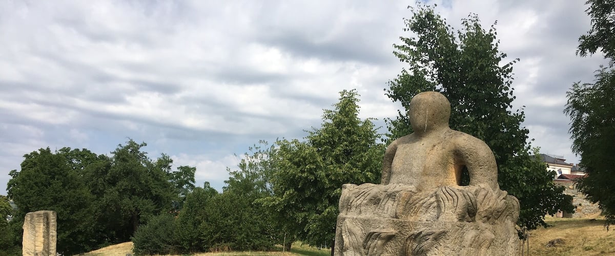 A stone statue of a seated figure with a lion's head on a pedestal in a grassy field with trees and a stone structure in the background.
