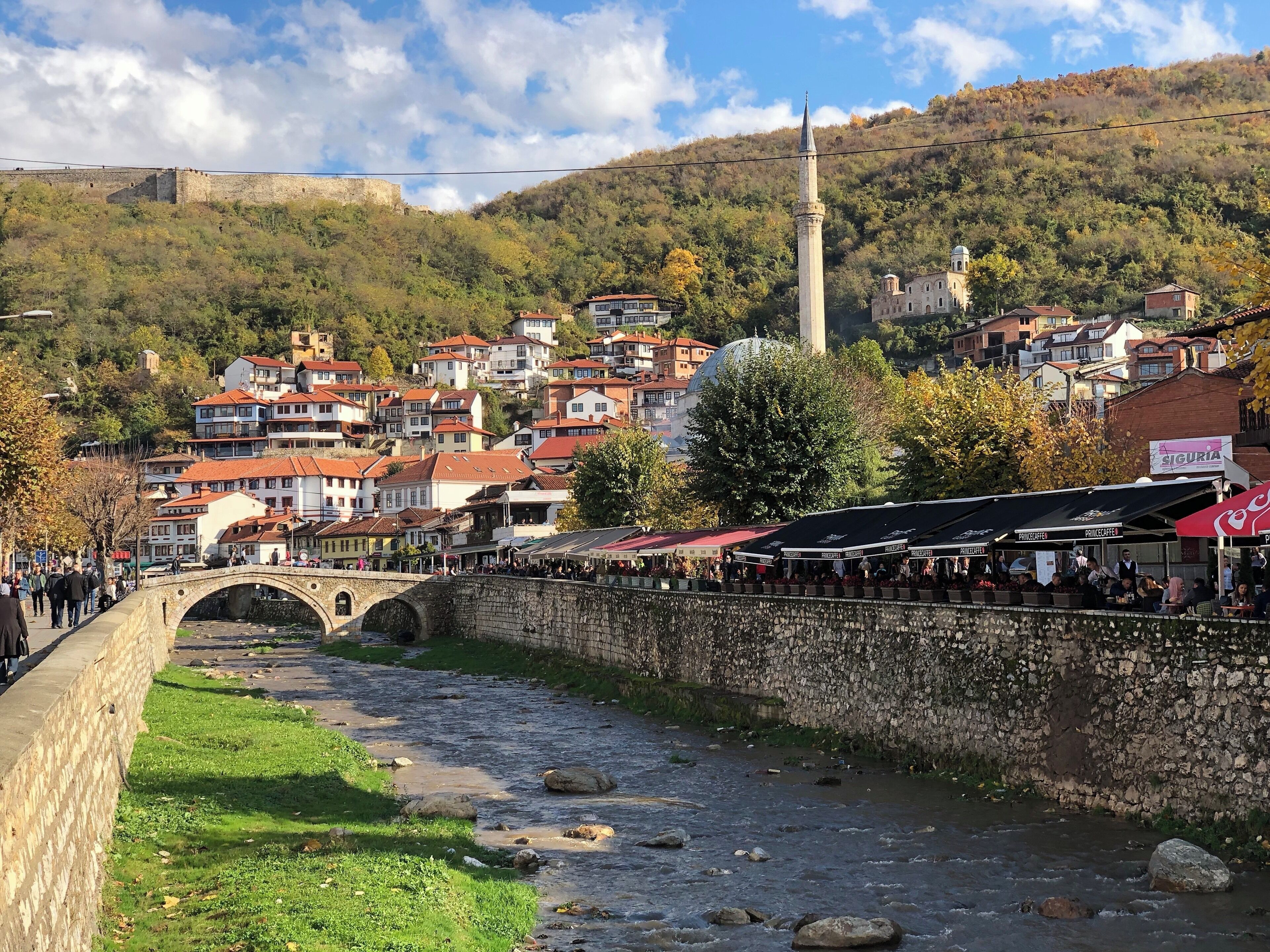 A river with a stone bridge, a mosque, and a hillside with buildings.