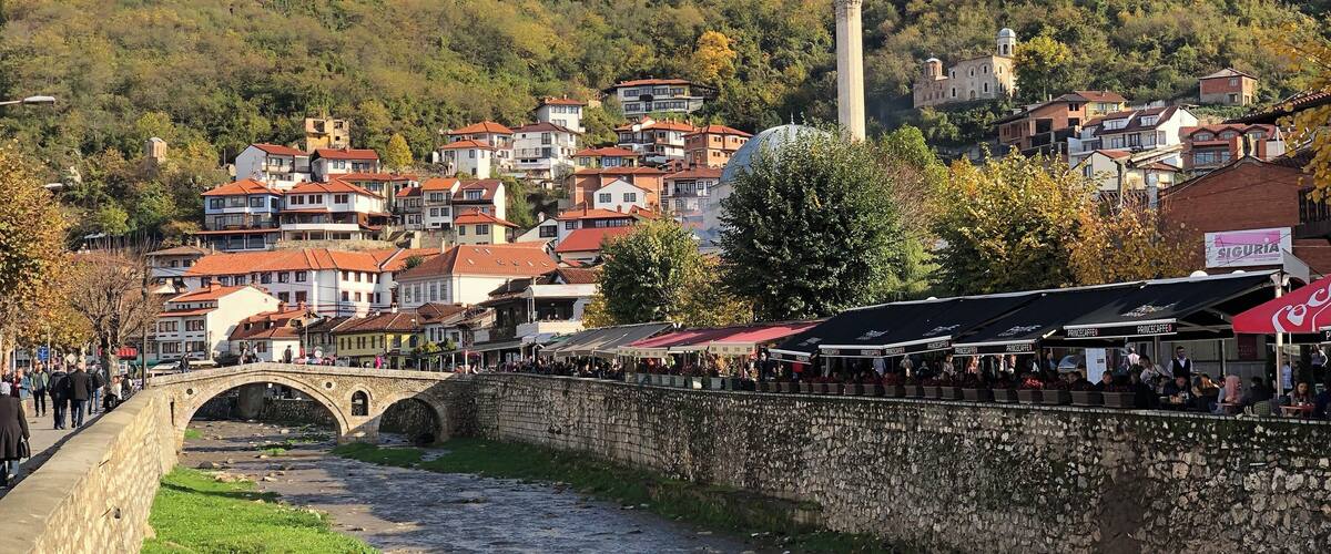A river with a stone bridge, a mosque, and a hillside with buildings.