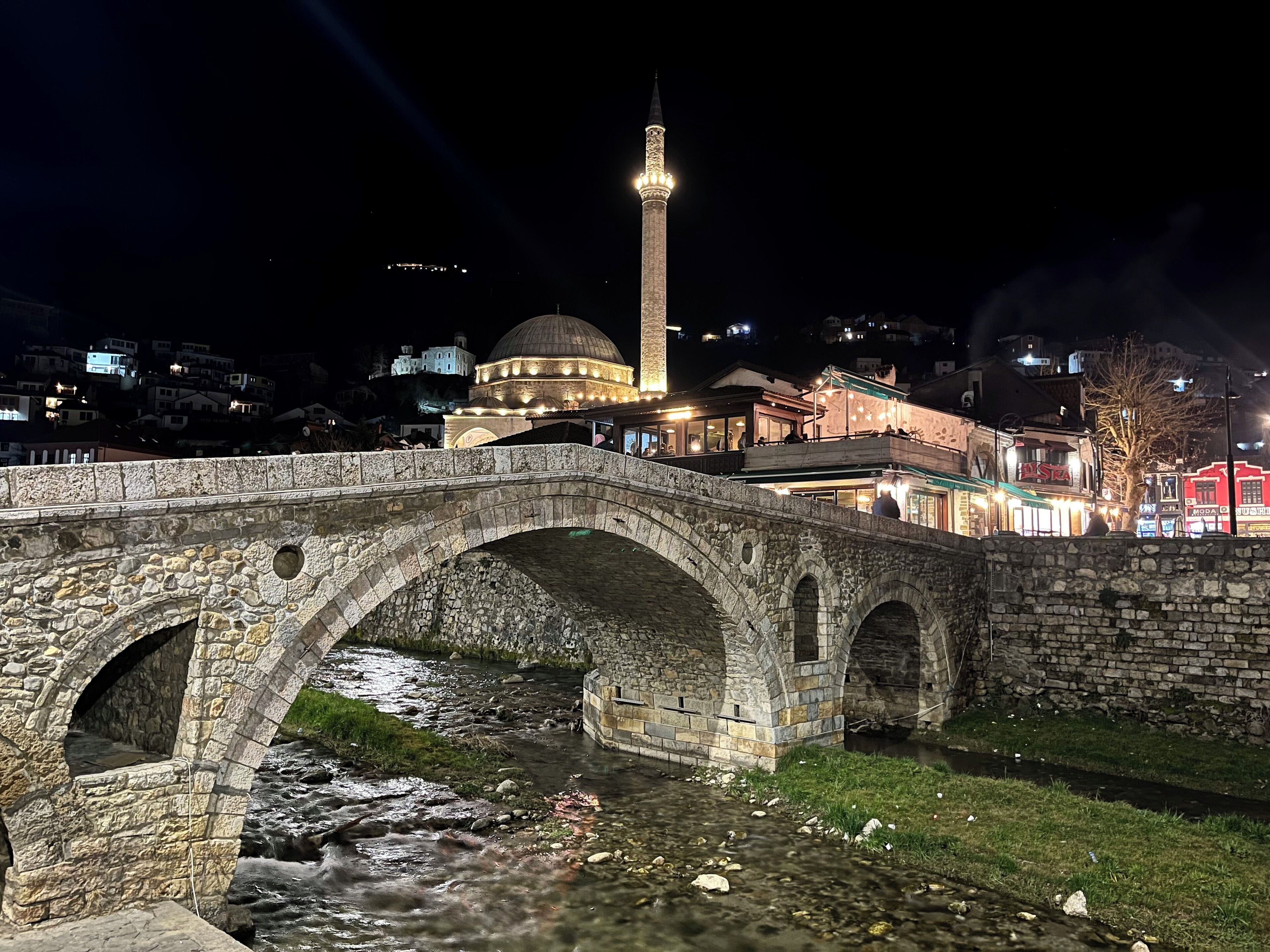 Un puente de piedra sobre un río, con una mezquita y un minarete al fondo.