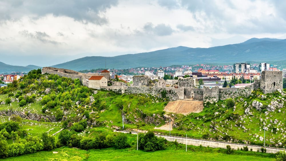 A historic castle on a hill overlooking a town with mountains in the background.